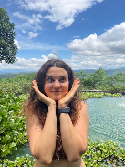 This media file is a portrait of a young woman taken outdoors. She is positioned in the foreground, facing the camera with her head tilted and resting on her hands, which are brought up to her face in a playful gesture. Her hair is long and curly brown, and she is wearing a dark watch on her left wrist. A faint red string is also visible on her wrist. Her expression is a soft smile, and her eyes are looking towards the camera.
The background is a vibrant and lush landscape. To the right of the woman, there is a body of water with some aquatic plants. Beyond the water, a green, hilly terrain stretches into the distance, with trees and vegetation covering the slopes. The sky above is a bright blue with scattered white clouds, suggesting it is daytime and the weather is pleasant. The overall scene evokes a sense of relaxation and enjoyment in a natural setting. The location context indicates this scene is in Sơn Điền, Vietnam. There is no visible text in the media file.