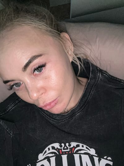 The image shows a person, likely female, with blonde hair, light skin, and striking blue eyes, looking directly at the camera. Her head is tilted to her left, suggesting she is lying down or reclined. She has prominent dark eyebrows and long, dark eyelashes.

She is wearing a dark, possibly charcoal-colored, distressed t-shirt. The shirt features a large white graphic design on the right side, which includes stylized letters, parts of which resemble "IN," along with other decorative elements that could be part of a logo or artistic motif. A small, faint tattoo is partially visible on her neck/shoulder area, beneath the collar of her shirt.

Her blonde hair is visible around her face and shoulder, resting against a light-colored pillow or fabric. The lighting in the scene is dim, indicating an indoor setting, possibly a bedroom, during the evening or night. The person's expression appears neutral or contemplative. This scene is located in Высокий, Russia.