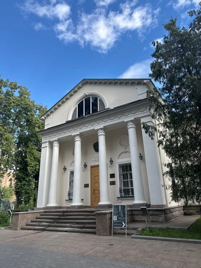 The image shows the exterior of a white classical-style building with large columns and a grand entrance. A semicircular window with black blinds is visible above the entrance. The building is surrounded by lush green trees and set against a bright blue sky with scattered white clouds, suggesting a sunny day. In the foreground, a paved walkway leads up a set of granite stairs to the building's entrance. A sign with an arrow and QR code is placed on the walkway. The scene appears to be a public building, possibly a museum or cultural institution, given the signage. The time of day appears to be daytime. The overall mood is serene and inviting, highlighting the architectural beauty of the building and its natural surroundings. The text on the sign is not clearly legible.