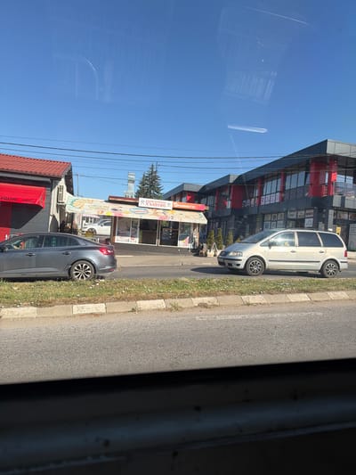 In Radomir, Bulgaria, on a bright, clear day, a photograph captures a roadside scene dominated by a clear blue sky. In the foreground, a grey sedan is parked on a grassy verge next to a paved road. To the right of the sedan, a light-colored minivan is in motion on the road, its interior showing occupants. Behind the vehicles, a row of buildings lines the street. One building, with a red roof and white exterior walls, features a red awning with Coca-Cola branding. Adjacent to it, a smaller building with a striped awning displays the text "БЪРЗО ХРАНЕНЕ ХЪШОВЕ" and "NESTLÉ". Further down the street, a modern, multi-story building with a black and red facade and large windows stands. Power lines crisscross the sky above the structures, and a tall, narrow metal structure is visible in the distance. The overall impression is of a commercial area along a road on a sunny day.