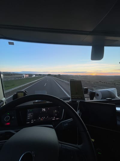 From the driver's seat of a vehicle, a highway stretches into the distance under a dawn or dusk sky. The dashboard displays a speed of 79 and a temperature of 26. A navigation device is mounted to the dashboard. Other vehicles are visible on the shoulder of the highway. A roll of paper towels sits on the dashboard. The steering wheel is in the foreground, partially obscuring the dashboard.