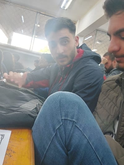 A selfie captured indoors showcases two men, one with closed eyes and a beard, positioned at the bottom edge. The other man, holding the camera, faces forward with a slightly puzzled expression. He is wearing a blue and red jacket. A person with a beard can be seen to the left of the two men in the foreground. The scene is filled with items and people, suggesting a crowded setting. A window with sunlight streaming through is in the background.
