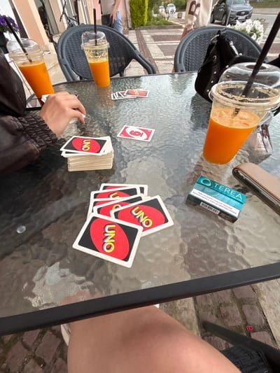 At an outdoor cafe in Garmen, Bulgaria, two clear plastic cups filled with orange juice sit on a glass-top table, each with a black straw. A stack of UNO cards is positioned to the left of the table, and other UNO cards are scattered in front of it. A hand, belonging to an unseen person, is visible reaching for the cards. To the right of the table, a pack of TEREA IQOS ILUMA cigarettes in a teal and blue package sits next to a brown leather case. In the background, people are visible, along with a parked car and a bicycle.