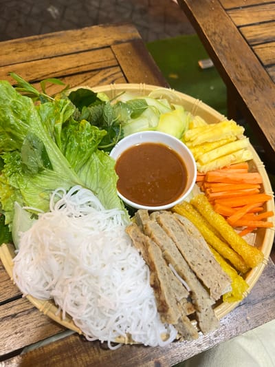 The image shows a platter of Vietnamese food, possibly a dish called Bún Chả, on a wooden table. The platter is round and appears to be made of woven material. It contains several components arranged around a central bowl of dipping sauce. These components include a large portion of white rice vermicelli noodles, fresh green lettuce leaves, various herbs such as mint, sliced cucumbers, julienned carrots, fried egg rolls or spring rolls, and grilled pork patties or sausage. The food is presented in a visually appealing manner. The background suggests an outdoor setting, with wooden planks and glimpses of green artificial turf, consistent with a restaurant or eatery in Bắc Hà, Vietnam. No people are visible in the image.