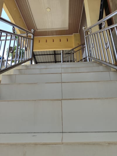The image shows a low-angle view of a tiled outdoor staircase leading upwards. The stairs are light-colored and appear to be made of large rectangular tiles. A polished metal handrail with decorative elements lines both sides of the staircase. The handrail is supported by vertical metal posts. Above the stairs, a yellow wall with small rectangular openings is visible, and beyond that, a white and brown paneled ceiling. The sky and some greenery, possibly palm trees, are visible in the background through the gaps in the railing. The scene is brightly lit, suggesting it is daytime. The location is identified as Uleemeuria, Indonesia.