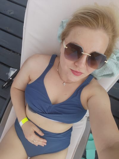 A woman lies on a lounge chair outdoors, on a wooden deck. She is wearing a blue two-piece swimsuit, large sunglasses, and a gold cross necklace. A yellow wristband is visible on her left wrist. Her hair is blonde and pulled back. The environment suggests a warm, sunny day, possibly at a resort or waterfront location in Pirogovo, Russia, given the proximity to reservoirs and dachas.