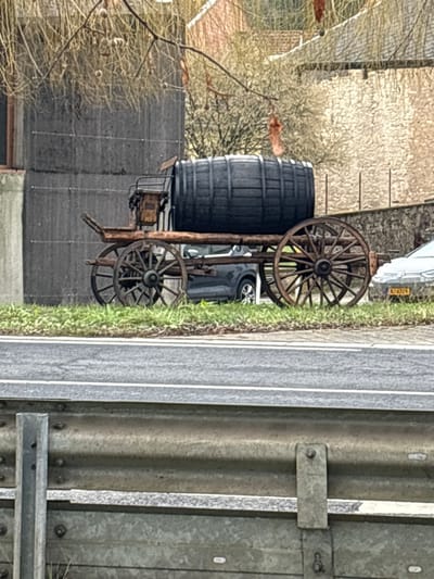 An antique-style wooden cart, featuring four large wooden spoked wheels, is positioned on a patch of grass beside an asphalt road in Bech-Kleinmacher, Luxembourg. A substantial, dark-stained wooden barrel rests horizontally on the cart's platform. A small, dark wooden structure is located at the front of the cart, above the front axle. Behind the cart, a dark grey modern car is partially visible, and further to the right, a lighter-colored car with Luxembourg license plate "NJ 4329" is also partially visible. The immediate foreground shows a grey metal guardrail above a section of the road with double white lines. In the background, to the left, is a tall, grey building structure, while to the right, a lighter-colored stone building with a reddish roof is discernible. Bare tree branches with some clinging foliage or seed pods extend across the upper portion of the frame, obscuring additional distant structures and trees. The scene appears under overcast lighting conditions. No individuals are present.