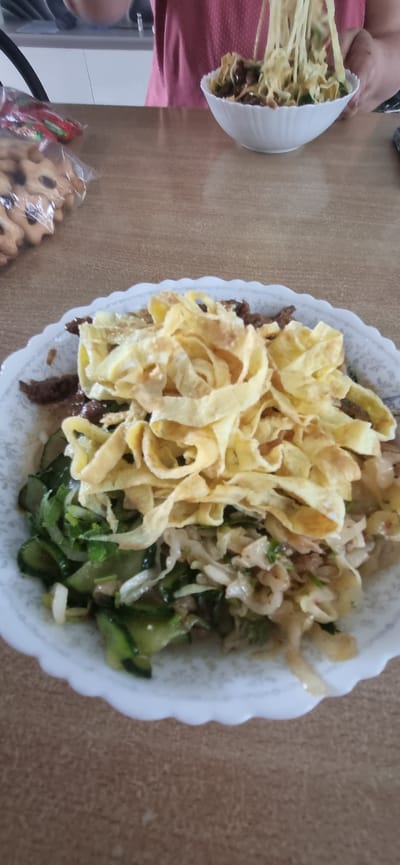 An overhead, eye-level shot captures a white scalloped bowl filled with a dish of noodles and vegetables, placed on a wood-grain table. The bowl is brimming with a layered mixture: at the bottom, sliced green cucumbers are mixed with what appears to be shredded cabbage and herbs. Layered on top of the vegetables are thin strips of fried egg, golden brown and piled high in the center of the bowl. A person's hands are visible in the background, holding a forkful of noodles, suggesting the dish is being prepared or served. To the left of the bowl, two plastic bags of cookies and a red packet are also on the table. The lighting is bright and natural, creating soft shadows. The background shows a sliver of a kitchen counter and a person wearing a pink t-shirt. The scene is likely indoors, possibly a dining area or kitchen in a home. The location is Blizhnee, Ukraine.