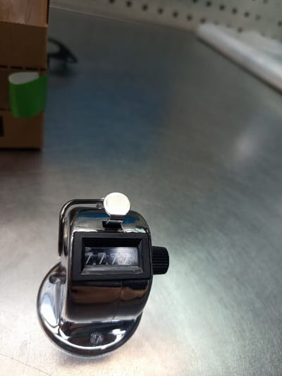 A chrome-plated manual clicker counter, displaying the numeric value "7777," rests on a brushed metal surface in an indoor setting. The counter features a silver actuation button on its top and a black knurled reset knob on its right side. In the upper left quadrant of the image, a segment of a brown cardboard box with a green adhesive strip is visible. The background further reveals a light-colored wall or surface with a pattern of dark dots, possibly a pegboard. This scene is located in Dettingen an der Erms, Germany.