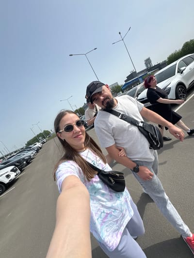 The photograph shows a group of people posing for a selfie outdoors, presumably in Bolshiye Kabany, Russia, though specific landmarks are not discernible. A woman holds the camera; she wears sunglasses and a patterned t-shirt. A man in a baseball cap and light t-shirt stands behind her. Another person, with reddish hair, appears to be lying on the hood of a white car in the background. Several cars are parked along the edge of the frame, suggesting a parking lot or roadside location. Streetlights are visible in the background. The overall impression is a casual, informal moment captured in a sunny outdoor setting.
