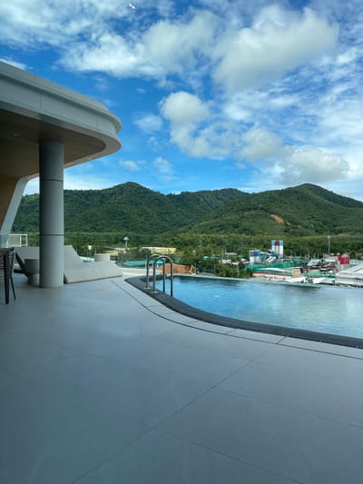 The image depicts a scenic view of a resort's infinity pool overlooking a lush, green mountainside with some construction happening in the distance. The sky is a vibrant blue with scattered white clouds, suggesting it's a clear day, likely in the late morning or early afternoon given the bright lighting.
The foreground features a modern, minimalist architectural element, possibly part of the resort's structure, with a curved design and a light-colored, textured finish. Adjacent to this is the edge of the infinity pool, with clear, blue water reflecting the sky. A set of silver pool ladders is visible, indicating the pool is ready for use. Beyond the pool, the scene transitions to a vibrant green landscape dominated by rolling hills covered in dense vegetation.
In the mid-ground, nestled within the hillside, are various structures indicative of ongoing construction. These include cranes, scaffolding, and buildings with colorful elements like red, white, and blue. There is also a tower-like structure visible further back. This suggests that the resort or a nearby development is either expanding or undergoing renovations.
A white sun lounger is placed near the pool, hinting at relaxation and leisure. A dark, modern chair and table are also visible, suggesting an outdoor seating area.
The location context of Si Sunthon, Thailand, combined with the tropical foliage and the resort-like amenities, places the scene in a warm, possibly humid climate. The overall mood is tranquil and inviting, despite the visible construction, conveying a sense of luxury and natural beauty. No discernible text is present in the image.