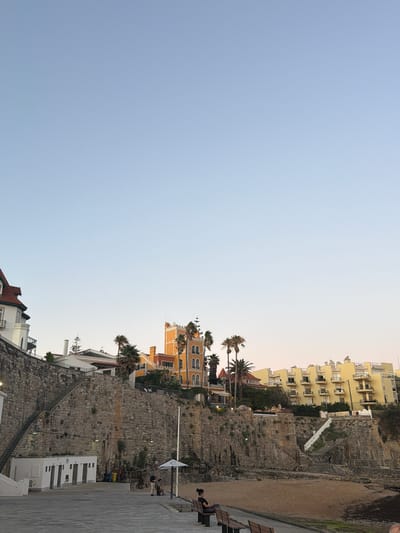This media file captures a scene in Cascais, Portugal, during what appears to be late afternoon or early evening, with a clear sky. The image is oriented sideways.
The dominant subjects are buildings, a rocky cliff face, and a paved walkway. On the right side of the image, several people are visible. Two individuals are seated on a park bench, with one appearing to relax. A few other people are seen walking or standing near the cliff base, some in the vicinity of a white building with a tiled roof.
The scene depicts a coastal urban environment. Buildings of various architectural styles, including modern apartment blocks with balconies and older, more ornate structures, are nestled along the elevated terrain. The cliff face is composed of rough-hewn stone, with vegetation growing in crevices and along its edges. A sandy area, possibly a small beach or embankment, is present at the base of the cliff. A white flag with a circular emblem is mounted on a pole on the cliff.
Notable details include the stonework of the cliff, the design of the buildings, and the presence of palm trees suggesting a warm climate. The lighting indicates a transition from daylight to dusk. The overall atmosphere seems peaceful and relaxed, with people enjoying the outdoor space. No visible text is present in the image.