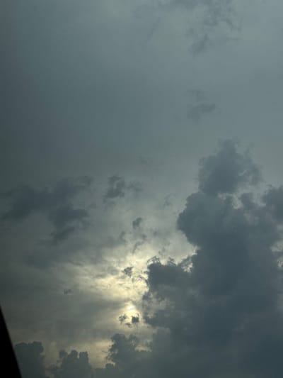 The image captures a dramatic sky filled with dark, heavy clouds. Sunlight pierces through gaps in the clouds, creating streaks of light and illuminating some of the cloud edges with a bright, almost golden hue. The overall mood is atmospheric and slightly ominous, suggesting an approaching storm or the aftermath of one. The clouds are varied in texture and density, ranging from wispy formations on the left to thick, brooding masses on the right. The interplay of light and shadow emphasizes the depth and turbulent nature of the sky. There are no discernible people, objects, or specific location cues, beyond the general impression of an outdoor, skyward view. The time of day is difficult to pinpoint precisely due to the cloud cover, but the quality of light suggests it could be late afternoon or early evening, when the sun is lower in the sky. No text is visible in the image.