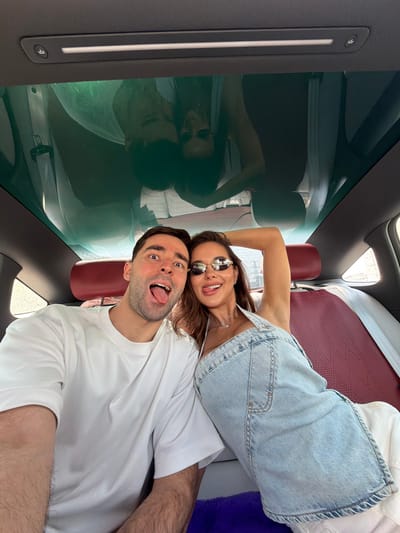 A selfie of a man and woman inside a vehicle in the Chaoyang District, China. The woman, wearing sunglasses and a white tank top with a light blue denim overall dress, sticks out her tongue. The man, wearing a white t-shirt, also sticks out his tongue. A clear glass roof reflects the scene. The interior features burgundy-colored seats. A control panel with a button featuring a person in a wheelchair is visible.