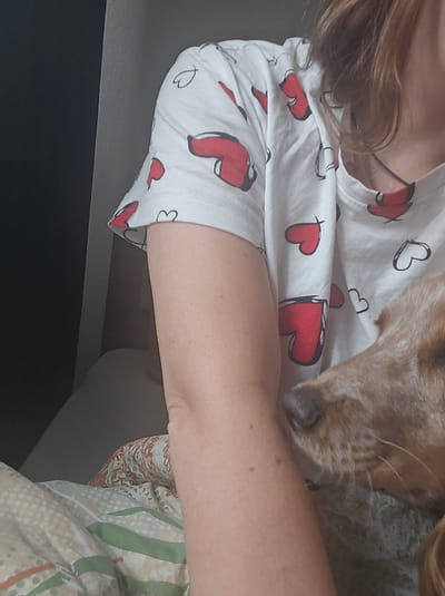 A close-up, waist-up shot shows a person's arm and shoulder wearing a white t-shirt with a red heart pattern. The person's arm is resting on a patterned blanket. A dog's head is visible in the foreground, its nose touching the person's arm. The background is dark and out of focus. The image appears to be taken indoors, possibly in a bedroom. There is no information available about the location in the city of Kimry, Russia.
