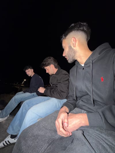 Three young men are posed closely together in an outdoor location in the city of Grad Rijeka, Croatia. The image is taken at night with the city lights visible in the background. The men are casually dressed in jeans and jackets/hoodies. The man at the top wears a black zip-up hoodie with a red logo, and gray sweatpants. His right hand is slightly injured. The other two men are wearing dark jackets and light-colored jeans. They seem to be standing on or near a stone ledge.