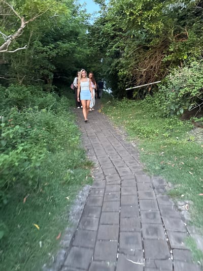 The image depicts a paved walkway lined with dense greenery, potentially in Uluwatu, Indonesia, given the tropical vegetation. Three individuals are walking along the path; the person in the foreground is fair-skinned with blonde hair, wearing a light-colored top and denim skirt. The individual behind them is fair-skinned with long red hair. The individuals appear to be on a leisurely stroll. The path is constructed of rectangular gray pavers. Vegetation, including trees and shrubs, borders both sides of the walkway, creating a tunnel-like effect.