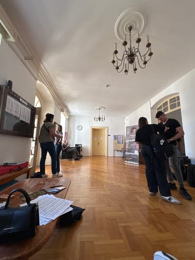 This is a candid, eye-level shot of the interior of a building, likely a public space such as a hallway or a communal area, possibly in Cetinje, Montenegro, given the poster visible in the background. The setting features polished wooden parquet flooring, high ceilings with ornate crown molding, and arched windows. A large, dark wood-framed bulletin board hangs on the left wall, displaying various documents.
In the foreground, a low, round wooden table is laden with sheet music, a black handbag, and what appears to be a book or folder. To the left of the table, a red bag rests on a wooden bench.
Several individuals are present in the scene. On the left, a person wearing a green t-shirt and jeans stands facing away from the camera, engaged in conversation with another person in a blue top. Further back, an older man is seated in a chair, looking at a device.
Towards the center of the image, a large poster with text and an image of a woman is displayed on the wall. To the right, two young people are interacting, one wearing a black t-shirt and the other a black top. The person in the black t-shirt is adjusting something on the other person's shoulder.
The lighting suggests it is daytime, with natural light streaming in from the windows and artificial light provided by a chandelier on the ceiling. The overall atmosphere appears relaxed and casual, with people engaged in conversation or preparing for an activity.