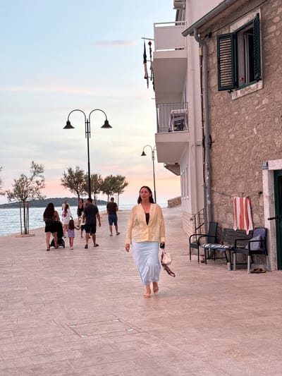 The image shows a woman walking on a paved waterfront promenade in Primošten, Croatia. The woman is wearing a light yellow blouse and a light blue skirt, and she is carrying a beige handbag. She is walking towards the viewer. In the background, several other people are walking along the promenade, some with children and a stroller. There are trees lining the promenade, and beyond them, the sea. The sky is a soft pink and orange, suggesting it is sunset. To the right of the woman is a building with a stone facade and a green shuttered window. There are benches and a towel hanging on the wall of the building. The street number "44" is visible above a doorway.