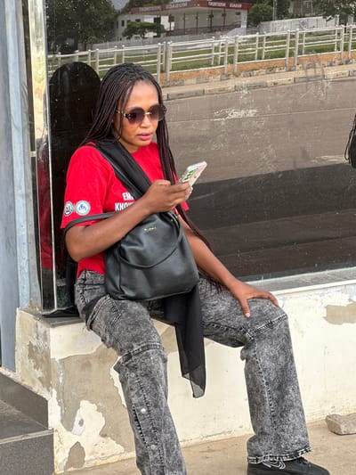 A Black woman wearing a red t-shirt and acid-washed jeans sits on a concrete ledge in front of a dark, reflective glass surface. She has braided hair and is wearing sunglasses. In her hands, she holds a mobile phone. A black crossbody bag rests on her lap. The background shows a street scene with a white fence, a road, and buildings, suggesting an urban environment in Unguwar Sarki, Nigeria. The concrete ledge has peeling paint.