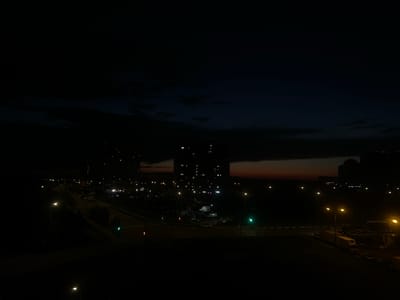 This media file depicts a nighttime urban scene in Domodedovo, Russia. The image is dark, with the sky transitioning from a deep blue to black, suggesting either dusk or pre-dawn hours. Wispy clouds are visible in the upper portion of the frame.
The main subjects are several tall apartment buildings, their windows lit, creating a grid pattern against the dark sky. Below the buildings, a road is visible, illuminated by streetlights and traffic signals. The road appears to be an intersection with at least two traffic lights, one showing green. Several vehicles, including what appear to be cars and a large truck, are present on or near the road, some parked in a lot.
The overall setting is a residential area with infrastructure typical of a suburban or city environment. The presence of illuminated buildings and vehicles indicates human activity, likely during a period of rest or commute. The mood conveyed is one of quietness and the ongoing life of a city at night. There is no visible text in the image.