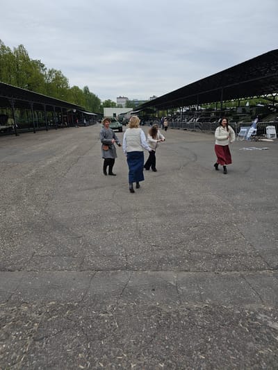 The scene depicts a paved area in what appears to be an open-air exhibition, likely located in Arkhangel’skoye, Russia, given the question's context.

Three distinct groups of people are present. The closest group consists of an older woman wearing a gray coat and hat, holding a handbag. Behind her is a mother with a child; the mother has blonde hair and wears a vest and blue skirt. The child is holding the mother's hand. Further back, another woman is visible, wearing a white jacket and a long red skirt.

To the left, a long, dark structure with a fence-like appearance stretches into the distance, possibly covering or bordering the exhibition area. Beyond this structure, trees with green foliage are visible. The sky is overcast with gray clouds. Vehicles and other structures can be seen in the far distance.