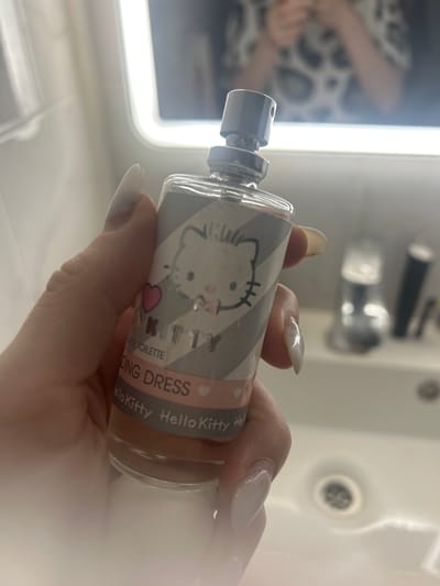 A person's hand with manicured nails holds a small bottle of Hello Kitty "Wedding Dress" Eau de Toilette. The bottle has a silver cap and a label featuring a graphic of Hello Kitty with grey and white stripes, and pink hearts. The background is blurred, showing a bathroom sink with a faucet and a mirror with a bright light. The image does not provide any information or context related to the city of Nizhniye Mull, Russia.