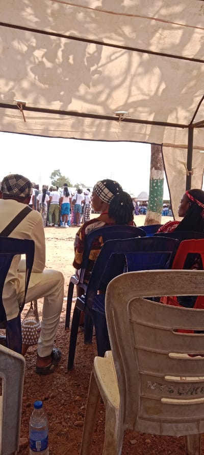 An outdoor scene in Ayere, Nigeria, depicts a gathering under a light-colored canvas canopy. In the immediate foreground, facing away from the camera, are three individuals seated on plastic chairs. On the left, a male figure wears a light beige top, light trousers, and a black and white patterned cap. A white beaded net bag, possibly a shekere, hangs from his side. A blue plastic water bottle rests on the reddish-brown, stony ground near his feet. Adjacent to him, a female figure with dark hair and a black and white patterned headband wears a multi-colored patterned dress. She is seated on a dark blue plastic chair. To her right, a partial view of a beige plastic chair is visible, with a hint of a red plastic chair beyond it.

In the midground, a crowd of approximately 20-30 individuals stands, predominantly facing away from the camera. Many are dressed in white tops, paired with various patterned skirts, wraps, or blue shorts. Their precise actions are not discernible beyond standing together. A light-colored tent structure is visible in the far background among this crowd. A tree with green and white paint on its lower trunk stands to the right of the standing group, supporting the canopy structure. Distant green trees are visible against a bright, hazy white sky, suggesting clear daytime conditions. Shadows of foliage are cast onto the underside of the canopy.