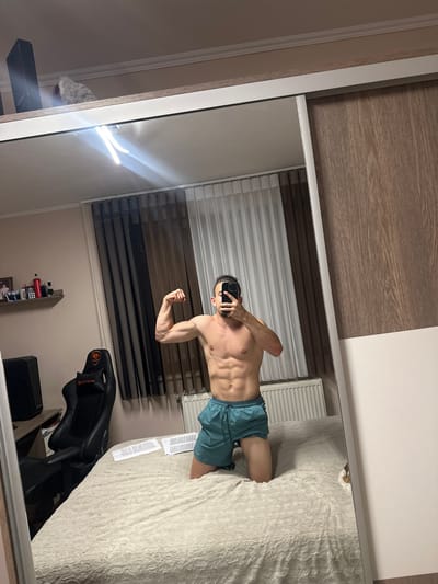 An indoor, vertical mirror selfie captures a male subject in what appears to be a bedroom located in the city of Saraia, Bulgaria.
The subject, a male with dark hair tied back in a bun and a beard, is shirtless, exposing a muscular torso with defined abdominal muscles. He is wearing teal-colored athletic shorts and is kneeling on a light-colored, fluffy bedspread. The subject is flexing his right bicep and holding a black smartphone directly in front of his face, obscuring it.
The room is reflected in a large, floor-to-ceiling mirror, likely part of a sliding wardrobe, which occupies the right half of the image. The wardrobe features a light wooden panel with a grain texture on the right side and a white frame with a silver sliding track. Above the wardrobe, a black electronic device and a white, fluffy object are visible on a ledge, beneath a light-colored ceiling with white crown molding.
To the left of the subject, also reflected in the mirror, is a beige-colored wall. A window is covered by white sheer curtains and dark brown opaque curtains. Below the window, a white radiator is installed. Further to the left, a black gaming chair with red accents and a "COUGAR" logo is positioned next to a dark-colored desk or table, on which a black speaker or computer tower rests. Above the desk, a wall-mounted shelf displays several items, including framed photographs, a red container, and a black bottle. Two sheets of paper with printed text are visible on the bedspread near the subject's left knee. The room is illuminated by artificial light, causing a strong glare to be reflected in the upper center of the mirror.