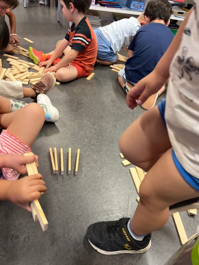The image depicts a group of young children playing with wooden blocks on a dark gray floor, likely indoors. In the foreground, a child's legs and feet are visible. They are wearing black athletic shoes with gold accents and a white and blue striped sock. Another child's legs are also visible, wearing a white sneaker and shorts.
Several wooden blocks are scattered on the floor. Some are laid out horizontally, resembling building blocks, while others are stacked vertically in a small group. One child, wearing an orange and black striped t-shirt and red shorts, is seated on the floor with blocks in front of them. Their right foot is wearing a bright orange and neon green sneaker. Another child, seen from the back, is wearing a dark blue t-shirt and light blue shorts, also appearing to be playing with blocks. A child in a white t-shirt and blue shorts is also present, leaning forward.
The environment suggests an indoor play area, possibly a classroom or playroom. The lighting appears to be artificial. The overall scene conveys a sense of children engaged in a constructive play activity. There is no information in the image to suggest a specific location in the city of La Fouillouse, France.