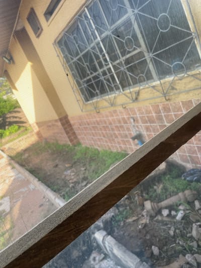 This image captures a view through a window screen, offering a glimpse of the environment outside. The foreground is dominated by a wooden beam and the fine mesh of the screen itself, which creates a textured overlay on the scene beyond.

Through the screen, we can see a courtyard area paved with light brown tiles arranged in a grid pattern. Adjacent to this tiled area, there appears to be a building with yellow walls and visible windows, some with horizontal louvers. Above one section of the building, a metal grate structure is visible, possibly part of a roof or an architectural feature.

Further into the background, the scene transitions to an unkempt outdoor area. This space is filled with loose debris, including stones, soil, and patches of green vegetation, suggesting a natural or slightly neglected environment. A light-colored pillar-like structure is also visible amongst the debris.

The lighting suggests it is daytime, with shadows indicating sunlight. The overall setting appears to be an outdoor space adjacent to a residential or utility area, possibly in a tropical or warm climate given the greenery. There is no discernible activity or people visible in the frame, and no text is present. The perspective is from an elevated viewpoint, looking down and across the scene.