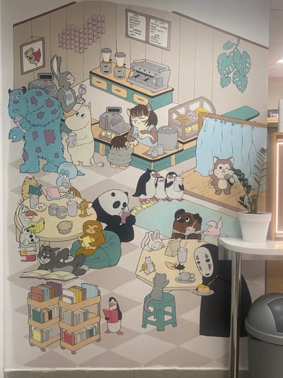 This image is a photograph of a mural painted on a wall in Kolomna, Russia, likely in a cafe or similar establishment, given the depiction of a coffee shop scene. The mural features numerous cartoon characters from various franchises, engaged in activities within a stylized coffee shop environment.
Prominent characters include a blue, furry monster resembling Sulley from Monsters, Inc., a white, Moomin-like character, a grey rabbit, a cat, penguins, a panda, a sloth, Olaf the snowman, Tom the cat, Jerry the mouse, and a character resembling No-Face from Spirited Away. The characters are shown ordering drinks, reading books, and socializing.
The coffee shop setting includes a counter with coffee machines and cash registers, shelves stocked with books, tables with food and drinks, and decorative elements like plants and a heart-shaped arrangement of pink squares on the wall. The overall aesthetic is whimsical and colorful, with a checkered floor pattern.
The photograph is taken from a slightly elevated angle, capturing a large portion of the mural. The lighting appears to be artificial, suggesting an indoor setting. In the foreground, a white table with a potted plant and a trash can are visible, indicating the mural is on a wall adjacent to a seating area.