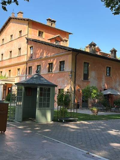 The image depicts an outdoor scene in front of a large, ochre-colored building, likely a hotel or villa, with several stories and numerous windows. The architecture suggests an Italian style, possibly with a historical feel. In the foreground, a small, dark green, octagonal structure with glass windows and a dark roof sits on a grassy area. Adjacent to this structure is a gold-colored luggage cart. Beyond these elements, there's a paved walkway leading towards the building. To the right of the luggage cart and in front of the building, an outdoor seating area with benches and an umbrella is visible. The ground is a mix of brick paving and grass. Sunlight casts shadows on the ground, indicating it is daytime. The overall setting appears to be well-maintained and tranquil. While the specific location in Fiumicino, Italy, is not explicitly stated in the visual cues, the architecture and ambiance are consistent with a setting in that region. There are no people visible in the frame.