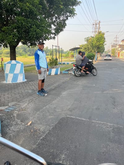 A man in a light blue long-sleeved shirt, grey shorts, and a black baseball cap is standing on a paved surface next to a blue and white checkered cylindrical object. He is smiling at the camera. Behind him, a motorcycle with two occupants, a man and a woman wearing a headscarf, is moving away from the camera. The scene appears to be a street in the city of Sugih Waras, Indonesia. There are trees and utility poles in the background, and the sky is clear. The ground is asphalt with some scattered debris.