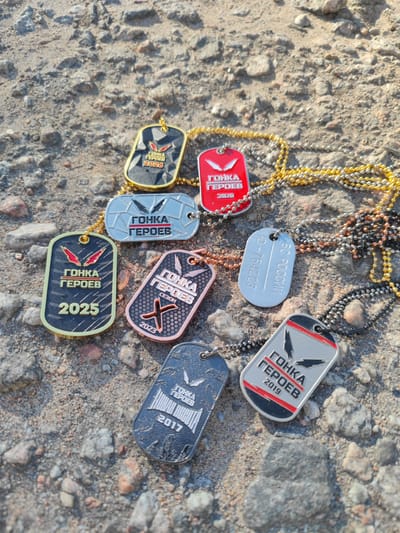 The image displays a collection of dog tags scattered on a gravelly, outdoor surface that appears to be a path or ground in Sertolovo, Russia. The dog tags are of various colors and designs, with some featuring text in Russian. One tag reads "ГОНКА ГЕРОЕВ 2026" (Race of Heroes 2026). Another says "ГОНКА ГЕРОЕВ 2020" (Race of Heroes 2020). A silver tag has "10-764223" and "ВС РОССИИ" (All of Russia) inscribed on it. A rose-gold colored tag reads "ГОНКА ГЕРОЕВ СЕЗОН 2023" (Race of Heroes Season 2023) with a stylized "X" symbol. A dark-colored tag displays "ГОНКА ГЕРОЕВ 2017" (Race of Heroes 2017). Another tag, predominantly green and black, is inscribed with "ГОНКА ГЕРОЕВ 2025" (Race of Heroes 2025). Finally, a grey and red tag reads "ГОНКА ГЕРОЕВ 2019" (Race of Heroes 2019). The dog tags are attached to various colored beaded chains: gold, silver, black, and copper. The background consists of rough, greyish gravel with some small stones. The lighting suggests it is daytime, with shadows cast on the ground. No individuals are present in the image.