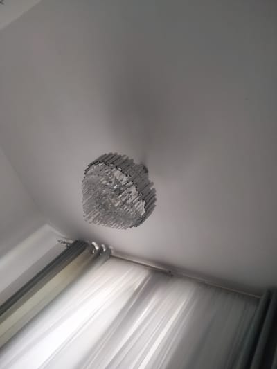 The image depicts a contemporary interior space, likely a room in a home. The primary object of focus is a modern chandelier suspended from a white ceiling. The chandelier has a unique design, appearing to be made of numerous small, rectangular crystal or glass elements arranged in a circular or heart-like pattern. It is illuminated, casting a soft glow.

Below the chandelier, a window is visible, adorned with two layers of curtains. The inner layer consists of sheer, white curtains that allow light to filter through, creating a soft, diffused illumination. The outer layer comprises thicker, darker curtains, possibly gray or a muted tone, which are partially drawn. The curtain rod is also visible.

The overall impression of the scene is one of modern elegance and domestic comfort. The lighting suggests it could be daytime, with natural light from the window complementing the chandelier's illumination. The setting appears clean and well-maintained, conveying a sense of order. There are no people visible in the image, and no text is present. The location context is Qaskeleñ, Kazakhstan.