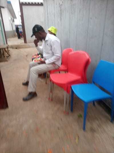 A man in a white shirt, light-colored pants, and a baseball cap sits on a red chair eating from a plate. Two more chairs, one red and one blue, stand beside him. Another person wearing a yellow hat is partially visible behind the man. They are situated outside, near a light gray wall and a building in the background. The ground is a light gray color with orange markings. The image is slightly blurred.