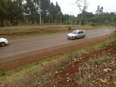 The image displays a two-lane asphalt road in Nkomo ward, Kenya, under an overcast sky. The road features double yellow center lines and is bordered by reddish-brown dirt shoulders and elevated banks with dry grass and sparse vegetation. A silver Toyota Probox-style van, fitted with roof racks, travels from right to left in the right lane. Behind and to the right of the van, an individual wearing dark clothing rides a black bicycle on the dirt shoulder, also moving from right to left. The front portion of a white car is visible on the far left, traveling in the opposite direction in the left lane. The background features a dense line of tall, slender trees, possibly eucalyptus, along with visible utility poles and a rudimentary wooden post fence running parallel to the road on an elevated bank.