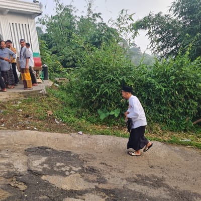 The scene takes place in a residential area of Kecamatan Tamansari, Indonesia, at the intersection of a paved road and a dirt path. A man wearing a black skullcap and a white shirt walks away from a group of men standing near a building. The man is carrying a black bag and wearing black pants. The men are wearing various clothing, including traditional Muslim attire. They appear to be speaking to each other.  The ground is covered in scattered leaves and debris. There is a building with a white facade and a white corrugated metal roof in the background. The building has a row of windows with metal bars. To the right of the scene, there is a dense thicket of bamboo. The sky is overcast, and there is a slight haze in the air. The location's latitude is -6.635, and longitude is 106.737.