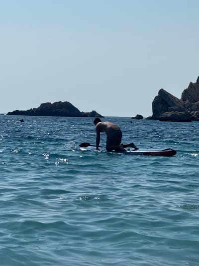 In the bright daylight, a person is paddleboarding in the azure waters off the coast of Perazića Do, Montenegro. The individual is kneeling on a blue and red paddleboard, facing away from the camera, and is positioned in the middle ground of the image. The water is choppy with sunlight reflecting off its surface. In the background, several rocky islets rise from the sea, with a clear blue sky above. The scene captures a moment of recreational activity in a coastal environment.