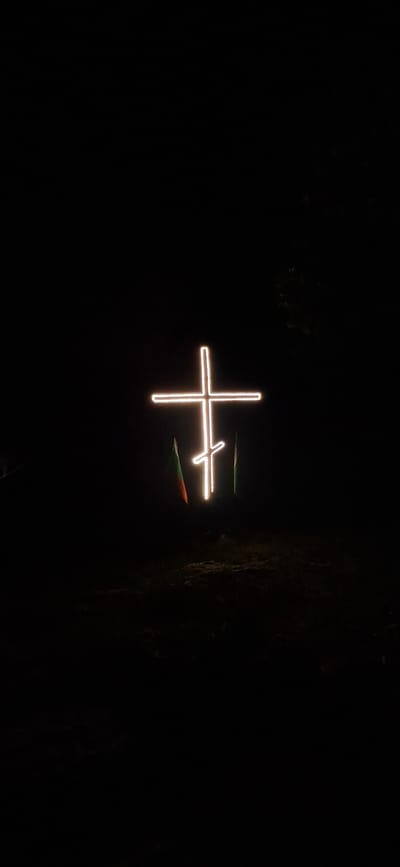 An outdoor nighttime photograph displays a prominently illuminated Eastern Orthodox cross, constructed from bright, white-yellow light strips, centrally positioned against a completely dark background. The cross features a main horizontal bar, a shorter horizontal bar positioned above the main one, and a lower diagonal bar slanted downwards from upper left to lower right. To the immediate left of the cross, a partially unfurled flag with visible stripes of green, white, and red is discernible, consistent with the national flag of Bulgaria, catching some light from the cross. To the immediate right, another flag is present, primarily exhibiting a vertical green stripe. The ground directly beneath the cross and flags is dark and uneven, suggesting natural, unpaved terrain. In the upper right background, faint, dark silhouettes of trees or foliage are barely visible against the dark sky. The scene is located in the city of Lyaskovets, Bulgaria.