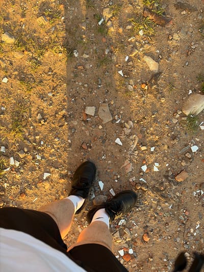 The image is an overhead view of a person standing on a dirt path littered with small stones, grass, and broken white debris. The person is wearing black shorts, white socks with a black logo, and black athletic shoes. The sunlight casts long shadows across the ground. The scene is not recognizable as being in the city of Syktyvkar, Russia.