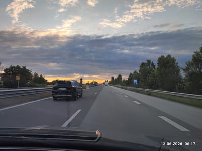 The image shows a view from a car traveling on a highway. The highway has multiple lanes, and the car is in the right lane. A black Kia SUV is in the left lane ahead of the car. The sky is cloudy with the sun setting, casting a warm glow on the clouds. Trees and vegetation line the sides of the highway. A road sign with international symbols is visible on the right side of the road. The timestamp at the bottom right of the image indicates June 13, 2026, at 06:17. The location is identified as the city of Tsaratsovo, Bulgaria.