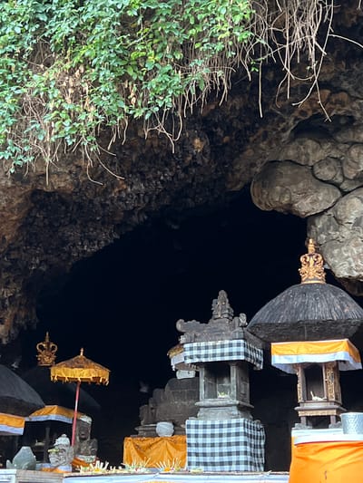 The image depicts the entrance to Goa Lawah Temple, a significant Hindu temple located in the village of Dawan, Klungkung Regency, Bali, Indonesia. The temple entrance is a dark cave opening, partially obscured by lush green foliage hanging from the rocky overhang above. In the foreground, several ornate Balinese structures are visible, adorned with black and white checkered cloth and bright orange fabric. These structures appear to be shrines or altars, characteristic of Balinese Hindu temples. One structure features a golden crown, while another has a golden umbrella with decorative tassels. The ground in front of the structures is covered with offerings, possibly consisting of flowers and incense. The overall scene suggests a place of worship, likely during a religious ceremony or a typical day at the temple. The lighting is dim, creating a mysterious and sacred atmosphere within the cave entrance and surrounding structures.