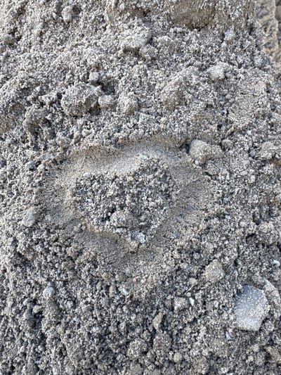 The image is a close-up, overhead shot of a patch of gray sand. The sand is composed of fine grains mixed with larger pebbles and clumps of compacted material. A prominent, crescent-shaped indentation is visible in the center of the frame, suggesting the imprint of a shoe or boot. The texture of the sand is rough and uneven, with shadows cast by the varied topography of the pile. The lighting indicates it is daytime, with sunlight illuminating the scene. There is no visible text. The image appears to be a neutral observation of a natural, outdoor surface, likely in a sandy or construction-related environment. Given the context of Usinsk, Russia, it could be a natural sand deposit or a construction site in the region.