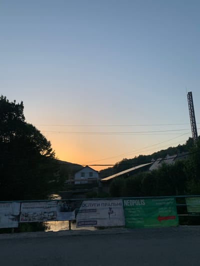 The image captures a serene sunset over the city of Sonyachne, Ukraine. The sky transitions from a pale blue at the top to a warm orange glow near the horizon, where the sun is setting behind a hill. Silhouetted against the vibrant sky are trees on the left and a cluster of buildings on the right, including a house with a white facade and dark-paned windows, and other structures with solar panels on their roofs. A tall, industrial-looking smokestack stands prominently on the right side of the frame. In the foreground, a river reflects the sunset's colors, and a bridge or barrier is visible. Along this barrier, several banners are displayed, advertising various services. One banner, green with white text, promotes "NEOPOLIS" and lists amenities like Wi-Fi, Smart-TV, a kitchen, air conditioning, and underfloor heating. Other banners advertise laundry services, business rentals, and massages. The overall scene is peaceful, with the fading light casting a tranquil ambiance over the urban landscape.