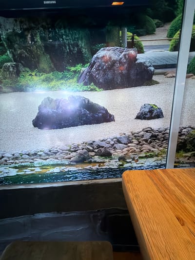 The image displays a digital artwork on a large screen, depicting a Japanese rock garden scene. The artwork features meticulously raked white sand, various sizes of rocks, lush green moss, and a small body of water with lily pads. In the background, there are carefully pruned bushes and a stone pathway with a wooden railing. The Samsung logo is visible at the top of the screen. In the foreground, a portion of a wooden table with a natural grain is visible on the right side. The overall ambiance suggests a serene and artistic setting. The image does not provide any information about the city of Donetsk, Ukraine, or any people, objects, or actions that would indicate its location.