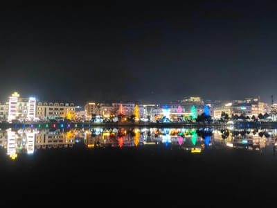 This image depicts a nighttime panoramic view of a section of Sa Pa, Vietnam, overlooking a body of water. The scene is dominated by a brightly illuminated cityscape reflected on the calm, dark surface of the water, which occupies the lower half of the frame.
Along the waterfront, a continuous row of multi-story buildings is visible, primarily constructed with light-colored facades and featuring numerous illuminated windows. Several of these structures display prominent signage, including "HA LOANG HOTEL," "SAPA PARADISE HOTEL," and "EYED HOTEL," indicating commercial or hospitality establishments. Interspersed among the buildings and lining the immediate edge of the water are numerous trees. Several of these trees are extensively decorated with multi-colored string lights, displaying hues of red, yellow, green, blue, and pink. A distinct row of circular, multi-colored lights (blue, green, red, yellow) is positioned directly at the water's edge, enhancing the festive illumination. The upper portion of the image shows a dark, cloudless night sky. No individuals or specific actions are discernible within the frame.