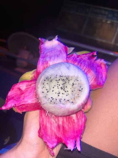 A human hand, consistent with a light skin tone, holds a partially peeled dragon fruit (Hylocereus undatus) in Lang Suan, Thailand. The fruit's exposed internal flesh is white, densely speckled with small, black edible seeds. The external peel is a vivid magenta, largely separated and pulled back from the fruit. The hand is positioned in the lower-left area of the frame, gripping the base of the fruit. The background is dark and out of focus, revealing a blurred circular object, possibly a fan or light fixture, in the upper-left, and a dark, indistinct surface to the right, possibly clothing or furniture. The scene is illuminated by a distinct bluish-purple light source.