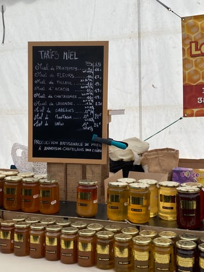 The image displays a honey market stall in Abondance, France, set up beneath a white canvas tent canopy. A prominent, vertically oriented blackboard sign, framed in wood, occupies the upper left-center, titled "TARIFS MIEL" (Honey Prices). The sign lists various honey types in white chalk, including "Miel de Printemps," "Miel de Fleurs," "Miel de Tilleul," "Miel d'Acacia," "Miel de Châtaigner," "Miel de Lavande," "Miel de Garrigues," "Miel de Montagne," and "Miel de Sapin," with prices provided for both 500g and 1kg quantities. Below the price list, the sign states "PRODUCTION ARTISANALE DE MIEL À ANNOISIN-CHATELANS DANS L'ISÈRE." A blue-handled clamp is attached to the right edge of the blackboard's support structure.
Below the sign, a tiered display table holds numerous uniform glass jars filled with honey. Each jar features a metallic golden screw-on lid and a rectangular golden label. Honey colors within the jars vary from light golden-yellow to rich amber and dark reddish-brown, reflecting different varieties such as "Miel de Lavande," "Miel d'Acacia," "Miel de Garrigues," and "Miel de Chataigner," as indicated on some labels. The display structure appears to be constructed from wooden crates or planks.
The background consists of the white canvas tent material, showing tension ropes and grommets. To the upper right, a portion of a banner with a honeycomb pattern and partially visible text, including "LO" and "38," is affixed to the tent wall. Behind the honey display, miscellaneous items are visible, including a crumpled light-colored cloth, a brown burlap sack, a cardboard box, and a purple package labeled "dolceuv." No individuals are present within the frame.